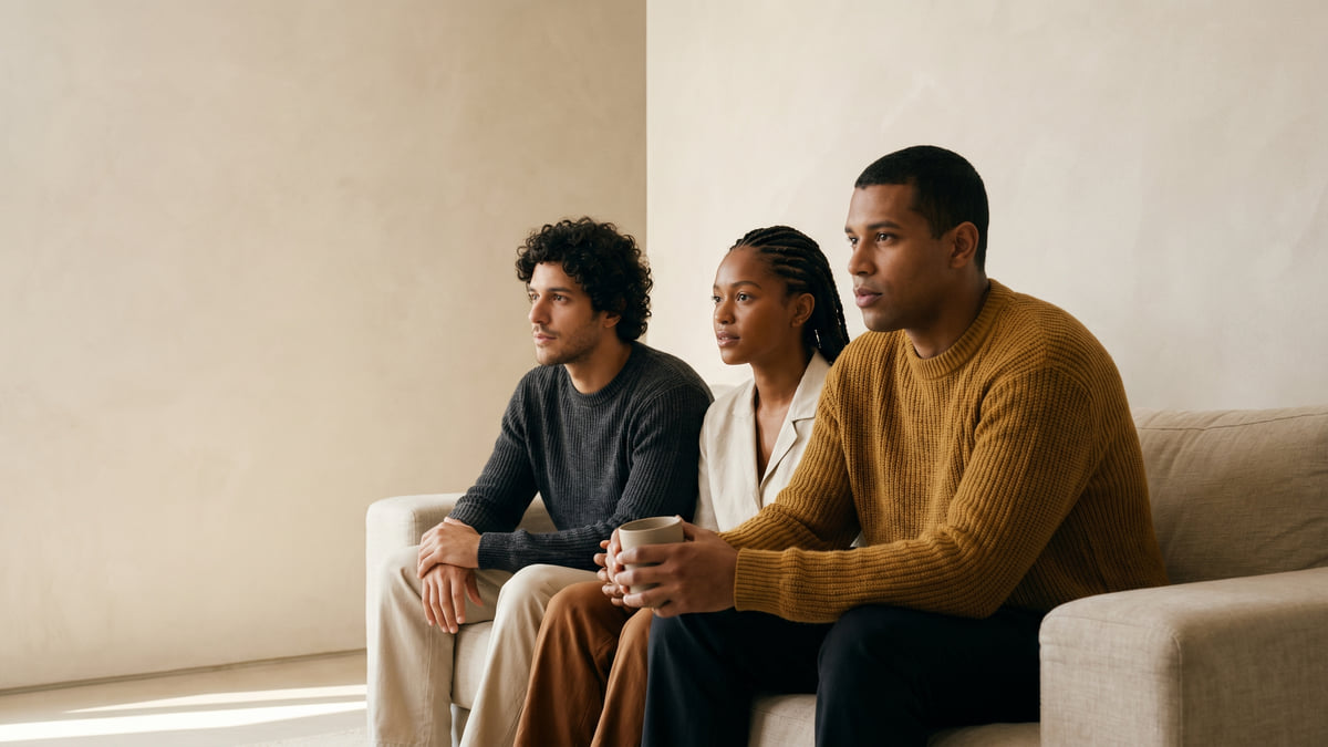 Três amigos reunidos num sofá assistindo a um jogo de futebol, em tons neutros e um suéter mostarda